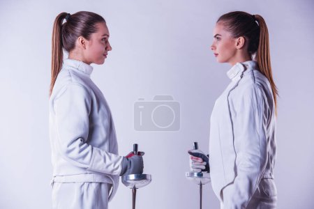 Photo for Beautiful female fencers in protective clothing are holding weapon and looking at each other, on gray background, side view - Royalty Free Image
