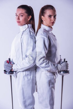 Foto de Hermosas esgrimistas femeninas en ropa protectora están sosteniendo el arma y mirando a la cámara mientras están de pie espalda con espalda sobre fondo gris, vista lateral - Imagen libre de derechos