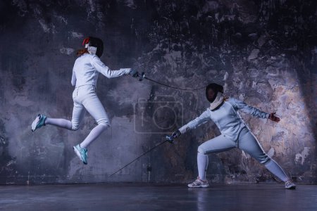 Photo for Female fencers in protective clothing are fighting on dark gray background, full-length - Royalty Free Image