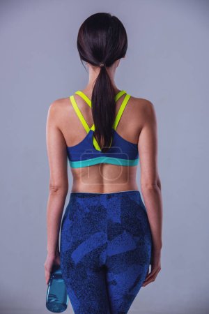 Foto de Vista trasera de chica atractiva en ropa deportiva sosteniendo una botella de agua, sobre fondo gris - Imagen libre de derechos