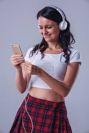 Attractive young woman in top and skirt is listening to music using a smartphone and smiling, on gray background