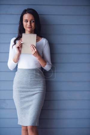 Photo for Attractive young woman is holding notepad and pen, thinking and smiling, on gray background - Royalty Free Image
