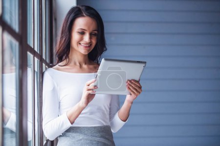 Téléchargez les photos : Attrayant jeune femme utilise une tablette numérique et souriant tout en se tenant debout dans la chambre - en image libre de droit
