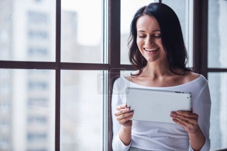 Attractive young woman is using a digital tablet and smiling while standing near the window