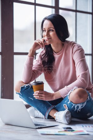 Téléchargez les photos : Jolie jeune femme en vêtements décontractés boit du café, regarde ailleurs et sourit tout en travaillant avec un ordinateur portable à la maison - en image libre de droit