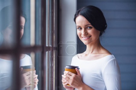 Téléchargez les photos : Jolie jeune femme tient une tasse de café, regardant la caméra et souriant tout en se tenant près de la fenêtre - en image libre de droit