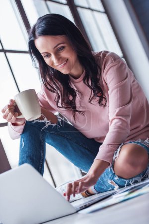 Téléchargez les photos : Attrayant jeune femme en vêtements décontractés tient une tasse et souriant tout en travaillant avec un ordinateur portable à la maison - en image libre de droit