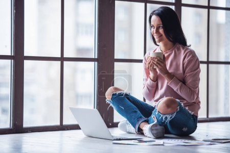 Foto de Mujer joven atractiva en ropa casual está sosteniendo una taza, mirando hacia otro lado y sonriendo mientras trabaja con un ordenador portátil en casa - Imagen libre de derechos