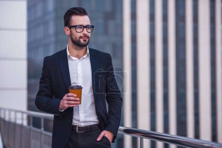 Foto de Hombre de negocios guapo en traje y vasos está sosteniendo una taza de café y mirando hacia otro lado mientras está de pie en el balcón - Imagen libre de derechos