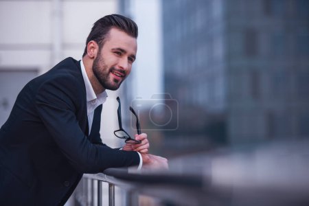 Foto de Hombre de negocios guapo en traje está sosteniendo gafas, mirando hacia otro lado y sonriendo mientras se inclina en el balcón - Imagen libre de derechos