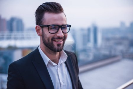Téléchargez les photos : Beau homme d'affaires en costume et lunettes sourit tout en se tenant sur le balcon - en image libre de droit