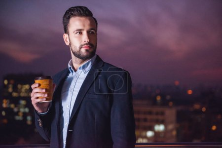 Foto de Hombre de negocios guapo en traje está sosteniendo una taza de café y mirando hacia otro lado mientras está de pie en el balcón por la noche - Imagen libre de derechos