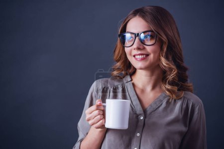Foto de Mujer joven atractiva en ropa casual y gafas está sosteniendo una taza, mirando hacia otro lado y sonriendo, sobre un fondo gris - Imagen libre de derechos