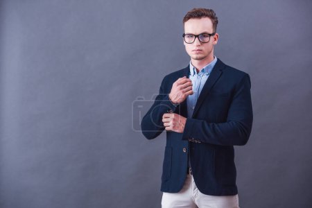 Foto de Guapo joven hombre de negocios en traje y gafas está ajustando gemelos y mirando a la cámara, sobre fondo gris - Imagen libre de derechos