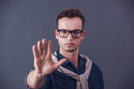Handsome young man in glasses is showing his palm and looking at camera, on gray background
