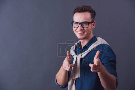 Téléchargez les photos : Beau jeune homme en lunettes pointe vers la caméra et souriant, sur fond gris - en image libre de droit