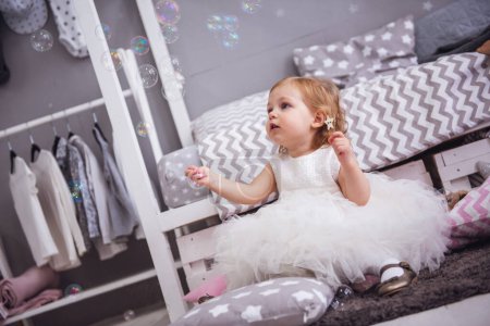 Téléchargez les photos : Petite fille mignonne en robe blanche moelleuse joue avec des bulles de savon dans la chambre des enfants - en image libre de droit