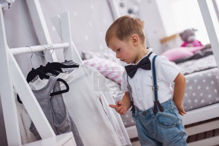 Foto de Lindo niño en jeans y pajarita está eligiendo ropa en la habitación de los niños - Imagen libre de derechos