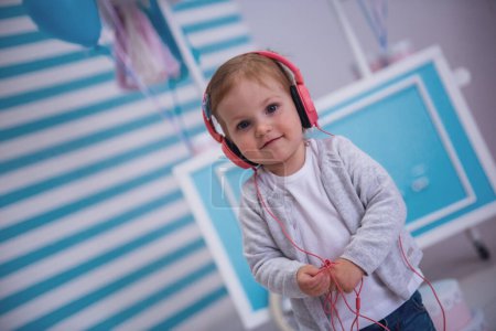 Téléchargez les photos : Petite fille mignonne dans les écouteurs regarde la caméra et sourire tout en écoutant de la musique dans la chambre des enfants - en image libre de droit