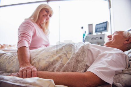 Téléchargez les photos : Vieille femme joyeuse regarde son mari couché dans le lit dans la salle d'hôpital, tenant sa main et souriant - en image libre de droit