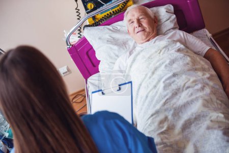 Téléchargez les photos : Beau vieil homme parle avec une infirmière et sourit tout en étant couché dans le lit dans la salle d'hôpital - en image libre de droit
