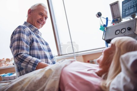 fröhlicher alter Mann blickt auf seine Frau, die auf der Krankenstation im Bett liegt und lächelt
