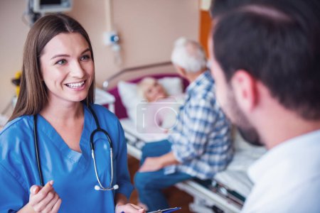 Photo for Old man is making a visit to his wife who is lying in bed in the hospital ward, nurse and doctor are talking in the foreground - Royalty Free Image