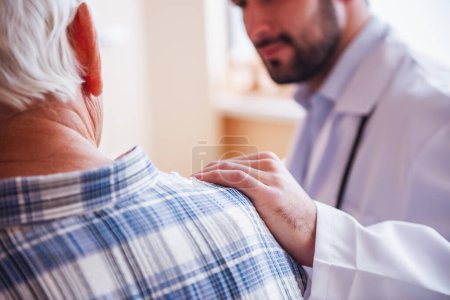 Foto de Imagen recortada de un médico guapo hablando con un anciano, sosteniendo la mano sobre su hombro y sonriendo mientras está sentado en la sala del hospital - Imagen libre de derechos