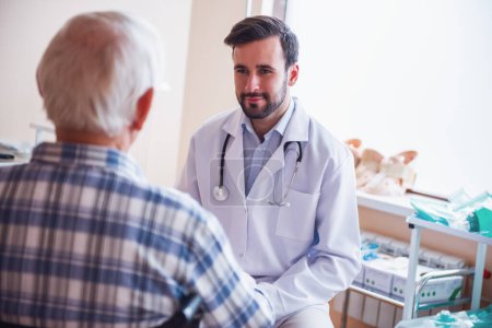Photo for Handsome doctor is talking with an old man in wheelchair in hospital ward - Royalty Free Image