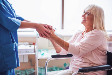 Foto de Hermosa anciana sostiene la mano del médico y sonríe mientras está sentada en la silla de ruedas en la sala del hospital - Imagen libre de derechos