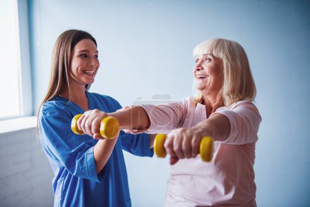 Foto de Hermosa anciana está haciendo ejercicios con pesas y sonriendo, en la sala del hospital. La atractiva enfermera la está ayudando. - Imagen libre de derechos