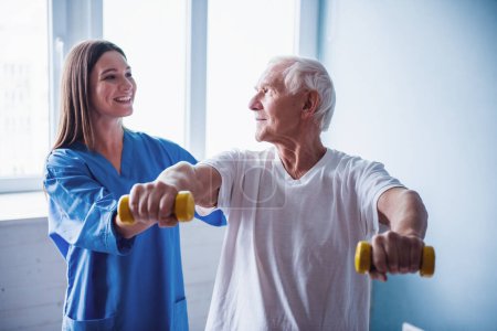 Foto de Un viejo guapo está haciendo ejercicios con pesas y sonriendo, en la sala del hospital. La atractiva enfermera lo está ayudando. - Imagen libre de derechos