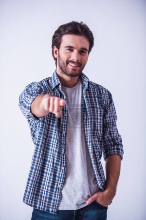 Photo for Handsome bearded man in casual clothes is pointing and looking at camera and smiling, on light background - Royalty Free Image