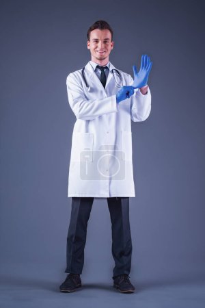 Foto de Imagen de cuerpo entero del guapo y joven doctor en bata médica poniéndose guantes, mirando a la cámara y sonriendo, sobre fondo gris - Imagen libre de derechos