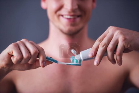 Foto de Imagen recortada de un joven guapo aplicando pasta de dientes sobre un cepillo de dientes y sonriendo, sobre un fondo gris - Imagen libre de derechos