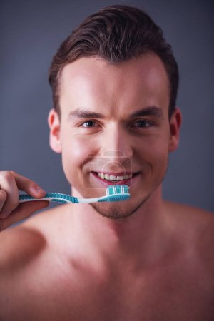 Téléchargez les photos : Portrait d'un beau jeune homme tenant une brosse à dents, regardant la caméra et souriant, sur fond gris - en image libre de droit