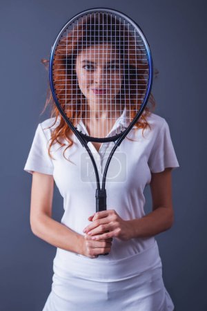Photo for Portrait of a young beautiful woman while playing tennis on a gray background holding a tennis racket in front of her face and a smile to the camera - Royalty Free Image