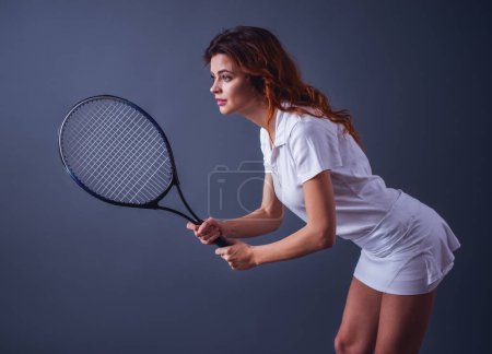 Photo for Portrait of a young beautiful woman while playing tennis on a gray background holding a tennis racket in front of her and a smile to the camera. - Royalty Free Image