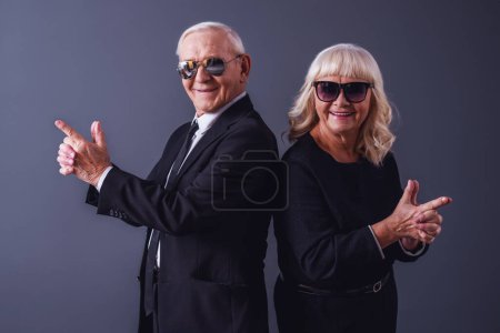 Photo for Beautiful old couple in elegant clothes and glasses is holding hands together imitating guns, looking at camera and smiling, on gray background - Royalty Free Image