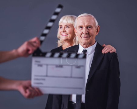 Photo for Beautiful old man and woman in elegant clothes on gray background, clapperboard in front - Royalty Free Image
