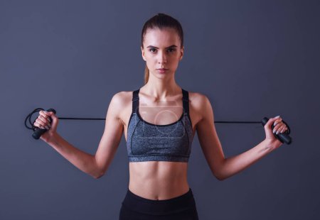 Photo for Beautiful girl in sportswear is holding a skipping rope and looking at camera, on gray background - Royalty Free Image