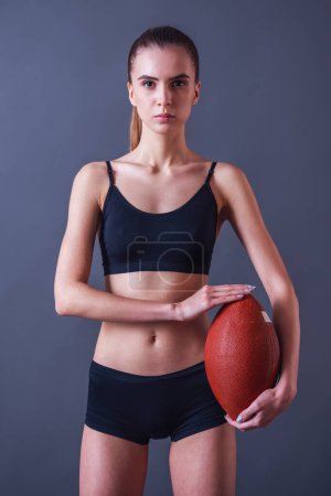 Photo for Beautiful girl in sportswear is holding an American football ball and looking at camera, on gray background - Royalty Free Image