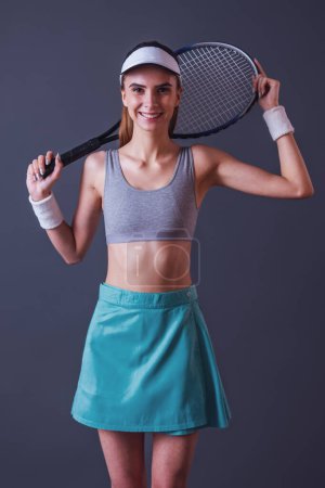 Téléchargez les photos : Belle fille en vêtements de sport tient une raquette de tennis, regardant la caméra et souriant, sur fond gris - en image libre de droit