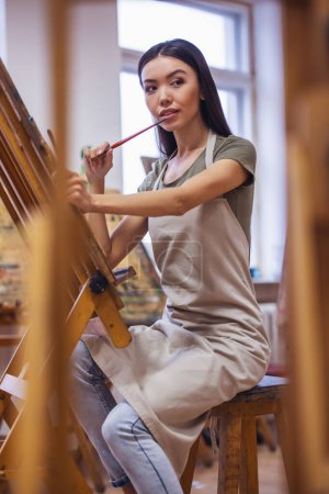 Foto de Joven hermosa mujer pintando artista mientras trabaja en un estudio, sonriendo a la cámara - Imagen libre de derechos