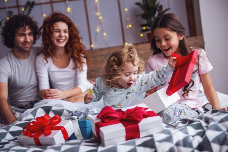Photo for It's Christmas time! Happy family sitting in bed, parents are smiling while their daughters are opening the presents - Royalty Free Image