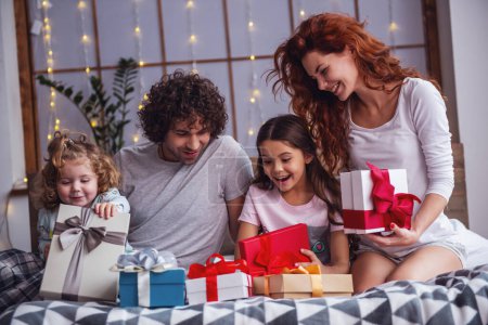 Téléchargez les photos : C'est Noël ! Heureuse famille assise au lit, les parents sourient pendant que leurs filles ouvrent les cadeaux - en image libre de droit
