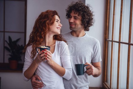 Téléchargez les photos : Beau couple boit du cacao chaud avec guimauve, se regardant et souriant tout en se reposant à la maison - en image libre de droit
