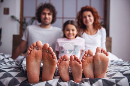 Téléchargez les photos : Les parents heureux et leur fille mignonne sourient tout en étant assis sur le lit, leurs pieds nus en évidence au premier plan - en image libre de droit