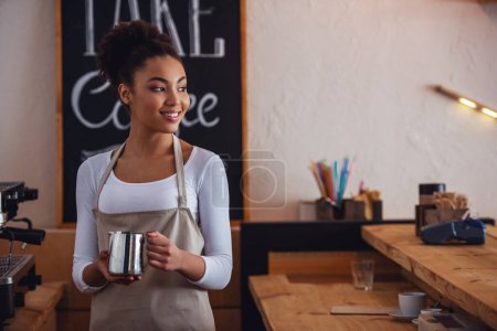 Hermosa barista afroamericana en delantal sostiene una taza de leche, mirando hacia otro lado y sonriendo mientras está de pie cerca de una máquina de café
