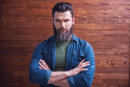 Photo for Serious bearded man in casual clothes is standing with crossed arms on a wooden background - Royalty Free Image